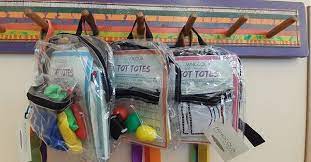 Three clear plastic backpacks hanging on a colorful wooden rack.