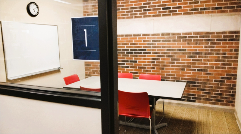 Study Room 1. A study room with a table, red chairs, brick walls, a whiteboard, and a clock and the number "1".
